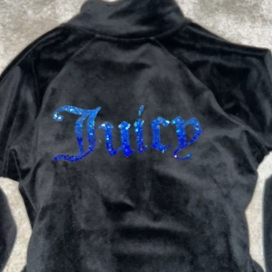 Svart velourjacka från Juicy Couture - Snygg svart velourjacka från Juicy Couture med glittrande blå text på ryggen. Jackan har en dragkedja framtill och en liten logga på bröstet. Perfekt för en avslappnad och trendig look.