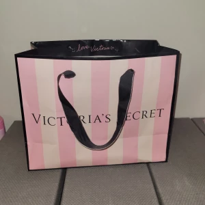 Rosa och svart påse från Victoria's Secret - Snygg påse från Victoria's Secret i rosa och svart med klassiska ränder. Väskan har svarta handtag och texten 'love, Victoria' på insidan. Perfekt för en shoppingtur eller som en stilren accessoar. Köparen står för frakten 