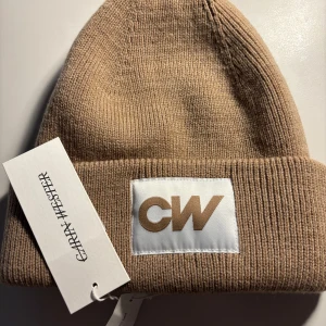 Beige ribbad mössa från Carin Wester - Snygg beige ribbad mössa från Carin Wester med CW-logga framtill. Perfekt för att hålla värmen under kyliga dagar. Stilren design som passar till det mesta.