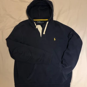 Mörkblå hoodie från Polo Ralph Lauren - Säljer en snygg mörkblå hoodie från Polo Ralph Lauren med det klassiska gula logotypen på bröstet. Hoodien har en knappdetalj vid halsen. Storlek är M och Nypris är (1500)