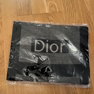 Svart och grå halsduk från Dior - Snygg och elegant halsduk från Dior i svart och grått med ett subtilt rutigt mönster. Perfekt för att ge en lyxig touch till din outfit. Halsduken är förpackad i plast och har en tydlig Dior-logga.