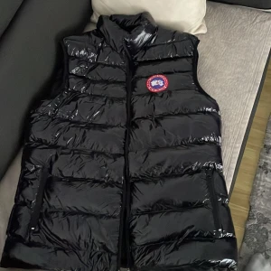 Svart dunväst från Canada Goose - Säljer en snygg svart dunväst från Canada Goose med glansig yta och dragkedja. Perfekt för kyliga dagar med sin stilrena design och varma fyllning. Västen har en hög krage och ett märke på bröstet.