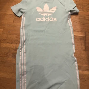 Ljusblå klänning från Adidas - Snygg ljusblå klänning från Adidas med korta ärmar och tryckt logotyp på ryggen. Klänningen har knappar längs sidorna och vita ränder som ger en sportig look. Perfekt för en avslappnad stil.