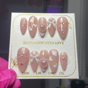 Handgjorda naglar med hjärtmönster och pärldetaljer - Säljer ett set av handgjorda naglar i en mjuk rosa nyans med vita hjärtan och pärldetaljer. Naglarna har en elegant design med rosetter och små stenar för extra glans. Perfekt för en romantisk look.