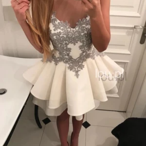 Vit klänning med grå spetsdetaljer - Elegant vit kortklänning med grå spetsbroderier på överdelen. Klänningen har en volangkjol och är ärmlös, vilket ger en festlig look. Perfekt för speciella tillfällen. Storlek S/36, från den polska hemsidan Lou.pl. Ordinarie pris 400zl (ca 1000 kr)