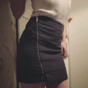 Grå kjol med dragkedja - Snygg grå kortkjol med en diagonal dragkedja framtill. Kjolen har en stilren design med dubbla lager vid fållen för extra detalj. Perfekt för en chic och modern.