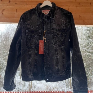 True Religion svart jeansjacka - Snygg svart jeansjacka från True Religion med klassisk design och knappar framtill. Jackan har två bröstfickor med lock och knapp samt en cool tvättad look. Perfekt för en avslappnad stil.