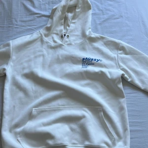 Stussy (jordgubbstryck) - Säljer en vit hoodie från Stüssy i storlek S. Den har en stor ficka fram och en cool jordgubbe tryckt på ryggen. På framsidan står det 'Stüssy' med flera städer listade under. Perfekt för en avslappnad och stilren look! Vid snabb affär 300kr