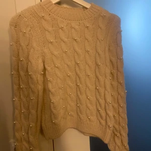 Beige stickad tröja med pärlor - Säljer en superfin beige stickad tröja med flätmönster och pärldetaljer. Tröjan är perfekt för höst och vinter, och den är både varm och mysig. Den har långa ärmar och en normal passform. Perfekt för att hålla sig stilren och varm under de kallare månaderna! 🧶✨