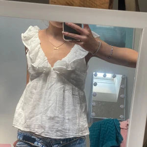 Vit blus med volangdetaljer - Säljer en superfin vit blus i storlek xs. Den har volangdetaljer vid axlarna och en v-ringad hals. Perfekt för sommaren eller en festlig tillställning. 