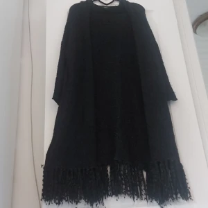 Svart lång kappa med fransar - Säljer en svart lång poncho med fransar längst ner, fickor på sidorna och en luva. Den är perfekt för höst och vinter och har en lös passform. Materialet känns mjukt och bekvämt, och den har långa ärmar. Perfekt för att hålla sig varm och snygg under de kallare månaderna.