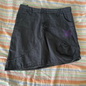 Svart kjol med broderi - Säljer en grå kjol i storlek 40. Kjolen har en cool design med sömmar som skapar ett mönster och ett lila broderi på framsidan. Den har fickor både fram och bak samt bälteshällor. 