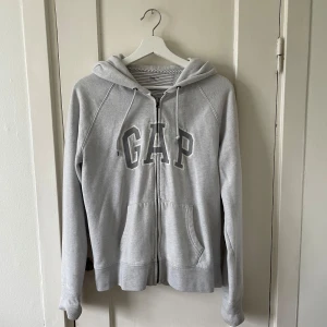 Grå hoodie från GAP - Säljer en grå hoodie från GAP i storlek L. Den är supermjuk och bekväm, perfekt för höst och vinter. Hoodien har en dragkedja framtill och två fickor. Den är i bra skick och har en klassisk GAP-logga på bröstet. 