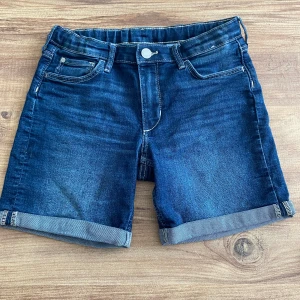 Mörkblå jeansshorts - Säljer ett par mörkblå jeansshorts i nyskick. De har en klassisk femficksdesign med knapp och dragkedja framtill. Shortsen har en snygg uppvikt kant nedtill som ger en avslappnad look. Perfekta för sommaren! 🌞