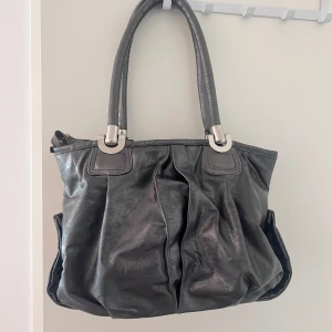 Grå/ silver handväska i skinn RETRO - Säljer en snygg handväska i skinn liknande material. Väskan har två robusta handtag och en dragkedja som kan vara lite problem med. Insidan är fodrad med brunt tyg och har en liten innerficka med dragkedja. Perfekt för både vardag och fest, väldigt stilren och trendig. 