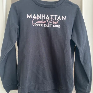 Svart tröja med Manhattan-tryck - Säljer en svart tröja med texten 'Manhattan Central Park Upper East Side' tryckt på framsidan i vitt och rosa. Tröjan är långärmad och har en normal passform. Perfekt för en casual look eller en mysig dag hemma. 🖤