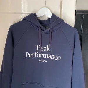 Peak Performance hoodie  - Säljer en snygg och bekväm blå hoodie från Peak Performance. Den har en klassisk design med loggan 'Peak Performance Est. 1986' tryckt på framsidan. Hoodien är i storlek XL och har en känguruficka samt justerbar huva med dragsko. Perfekt för både va