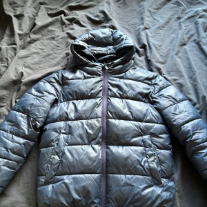 Svart pufferjacka från Hugo Boss - Säljer en snygg svart pufferjacka från Hugo Boss. Jackan har en quiltad design med en praktisk huva och dragkedja framtill. Perfekt för kalla dagar och håller dig varm och stilren. Den är i nyskick och redo för nya äventyr!