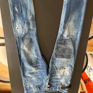 Säljer ett par coola blå jeans från DSQUARED2. De har en distressed look med flera slitningar och en stor lapp på ena benet. Jeansen har en klassisk femficksdesign och en röd DSQUARED2-logga på baksidan. Nypris 6500kr