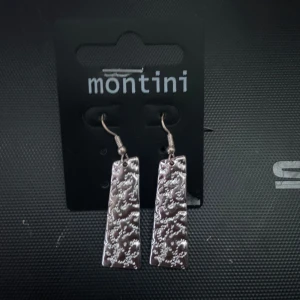 Silvriga örhängen från Montini - Snygga och stilrena silvriga örhängen från Montini. De har en rektangulär form med ett unikt mönster som ger en elegant touch. Perfekta för både vardag och fest!