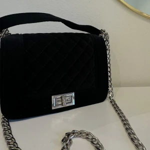 Elegant svart axelväska med kedjedetalj - Snygg och stilren svart axelväska med quiltat mönster och silverfärgad kedja. Väskan har en elegant låsdetalj i metall och passar både till vardags och fest. Väldigt lättanvänd och i fint skick! 