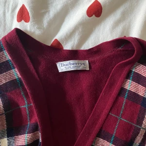 Rutig kofta från Burberrys - Säljer en superfin rutig kofta från Burberry i en djup vinröd färg. 100% Pure lambwool!  Den har ett klassiskt rutmönster i beige, blått och grönt. Koftan är långärmad och har knappar framtill. Perfekt för höst och vinter när du vill vara både stilren och varm!