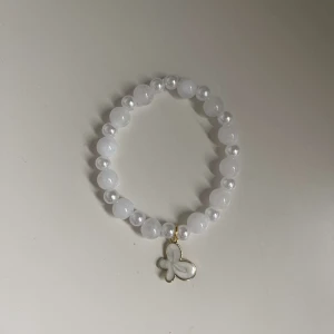 Vitt pärlarmband med fjärilsberlock - Säljer ett superfint vitt armband med pärlor och en söt fjärilsberlock i guld. Perfekt för att ge en elegant touch till vilken outfit som helst. Armbandet är elastiskt och passar de flesta handleder. Perfekt för både vardag och fest! 🦋✨