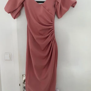 Rosa klänning med puffärmar - Säljer en superfin rosa klänning med puffiga kortärmar och en elegant drapering på sidan. Klänningen har en dragkedja i ryggen och är perfekt för festliga tillfällen. Den är i nyskick och har en vacker passform som framhäver figuren. Den har prislappen kvar, storlek 32. PRIS KAN DISKUTERAS!