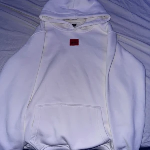Vit hoodie från Hugo Boss - Säljer en stilren vit hoodie från Hugo Boss. Den har en röd broderad logga på bröstet och en klassisk känguruficka framtill. Perfekt för en avslappnad och trendig look. Passar både till vardags och chillkvällar. 🕶️