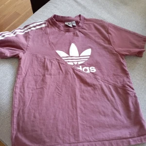 Rosa t-shirt från Adidas - Säljer en snygg rosa t-shirt från Adidas med det klassiska treklöver-loggan i vitt på framsidan. T-shirten har korta ärmar och de ikoniska tre ränderna på axlarna. Perfekt för en avslappnad stil eller träning. Materialet känns mjukt och bekvämt.