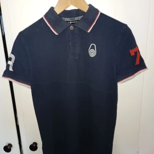 Mörkblå pikétröja från Sail Racing - Säljer en snygg mörkblå pikétröja från Sail Racing. Den har korta ärmar och en klassisk krage med röd och vit kant. På ena ärmen finns en röd siffra 7 och på den andra en vit siffra. Perfekt för en sportig look!