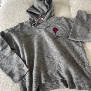 Grå hoodie med rosdetalj - Säljer en supermysig grå hoodie från Koton i storlek L. Den har en snygg röd ros broderad på bröstet och en klassisk huva med dragsko. Perfekt för kyliga dagar när du vill vara både bekväm och stilren. Passar perfekt till jeans eller leggings!