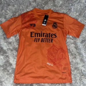 Orange fotbollströja från Adidas - Säljer en snygg orange fotbollströja från Adidas med korta ärmar. Tröjan har tryck med 'Emirates Fly Better' och Real Madrids logga på framsidan. Den är i nyskick och kommer i originalförpackning. Perfekt för fotbollsfans!