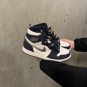 Nike Air Jordan 1 Japan Midnight - Snygga Nike Air Jordans i en stilren kombination av blått och vitt. Skorna har en klassisk design med snörning och en silverfärgad Nike-logga på sidan. Perfekta för dig som vill ha en ikonisk stil.