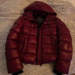 Vinröd jacka från Superdry - Säljer en snygg vinröd jacka från Superdry. Jackan har en huva och stängs med både dragkedja och knappar. Perfekt för kyliga dagar med sin vadderade design.