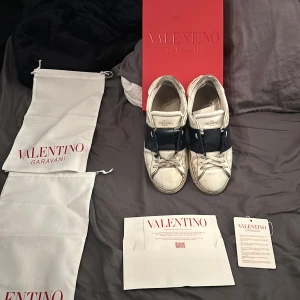 Valentino Garavani sneakers - Snygga Valentino Garavani sneakers i vitt skinn med en bred mörkblå rem över ovansidan. Skorna har en klassisk design med snörning och en bekväm passform. Perfekta för en stilren look. Skicket är inte det bästa pris kan diskuteras 