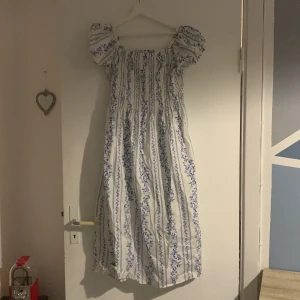 Vit och blå blommig klänning - Charmig vit klänning med blått blommönster och puffiga kortärmar. Klänningen har en romantisk stil med en luftig passform och elastisk halsringning.