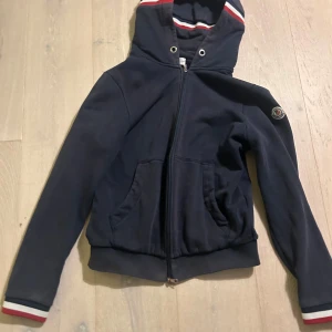 Mörkblå zip-hoodie från Moncler - Säljer en snygg mörkblå zip-hoodie från Moncler. Tröjan har en broderad logga på ärmen och röd-vita detaljer vid huva och ärmslut. Finns små defekter som såns på bild, samt bild på monclerscan och bild på moncler koden som kan verifieras på monclercode hemsidan! Perfekt för en stilren look! Nypris 5000, men modellen säljs inte längre.