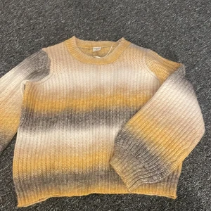 Randig stickad tröja i gult och grått - Mysig stickad tröja med ett randigt mönster i gult, grått och beige. Tröjan har långa ärmar och en rund halsringning. Perfekt för kyliga dagar.