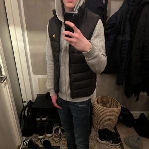Svart dunväst från lyle & scott  - Snygg svart dunväst med en gul logga på bröstet. Västen har en dragkedja och är perfekt för lager-på-lager under kyligare dagar. Den är stilren och lätt att matcha med olika outfits.