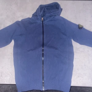 Blå huvtröja från Stone Island - Säljer en snygg blå huvtröja från Stone Island med dragkedja och ribbade muddar. Tröjan har en broderad logotyp på ärmen och är perfekt för en casual look. Köpt på mytheresa och är 100% äkta. Nypris 3800kr