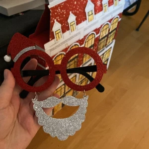 Julglasögon med tomteluva och skägg - Roliga julglasögon med runda röda bågar och en fastsatt glittrig tomteluva och skägg. Perfekta för att sprida julstämning på festen!