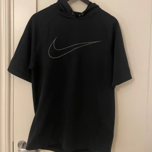 Svart kortärmad hoodie från Nike - Säljer en svart kortärmad hoodie från Nike med den ikoniska loggan på bröstet. Hoodien är tillverkad i en bomulls- och polyesterblandning, vilket gör den både bekväm och hållbar. Perfekt för en avslappnad stil.
