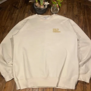 Beige sweatshirt från H&M - Säljer en beige sweatshirt från H&M med texten 'STAY IN SLOW MOTION ESCALATE IN SPACE' i guld på både fram- och baksidan. Tröjan har en relaxed fit och långa ärmar, perfekt för en avslappnad stil.