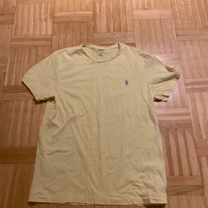 Ljus Gul t-shirt från Ralph Lauren - Säljer en ljusgul t-shirt från Ralph Lauren med en broderad blå logga på bröstet. T-shirten är kortärmad och tillverkad i bomull, perfekt för en avslappnad stil.