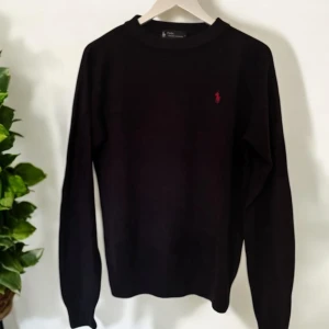 Svart tröja från Polo Ralph Lauren - Snygg svart tröja från Polo Ralph Lauren med en liten röd logga på bröstet. Tröjan har en klassisk rund hals och långa ärmar, perfekt för en stilren look.