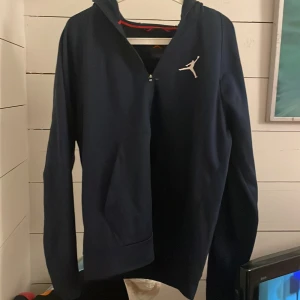 Mörkblå hoodie från Jordan - Säljer en snygg mörkblå hoodie från Jordan med dragkedja och det ikoniska logotyptrycket på bröstet. Perfekt för en sportig look eller avslappnade dagar. Hoodien har långa ärmar och en praktisk huva.