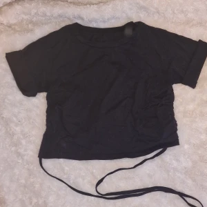 Svart t-shirt med snörning - Svart tajt/baby tee med lite kortare rygg och snörning baktill från sidorna. Oklart vilket märke, finns ingen lapp. Knappt använd.