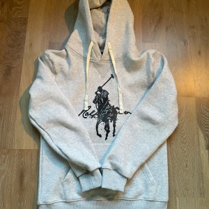 Grå hoodie från Polo Ralph Lauren - Snygg grå hoodie från Ralph Lauren med ett stort tryck av Polologgan på framsidan. Perfekt för en avslappnad stil. Tröjan är Storlek M men passar mest S. Modellen är 171cm och 65kg. Hör av dig vid minsta fundering!