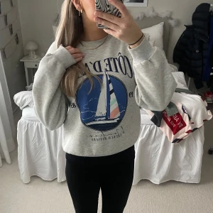Grå sweatshirt med segelbåtsmotiv - Säljer en grå sweatshirt med ett stort tryck av en segelbåt och texten 'Côte d'Azur'. Tröjan har en rund hals och långa ärmar, perfekt för en avslappnad stil. Den är mjuk och bekväm, idealisk för kyligare dagar. Väldigt fint skick då den knappt är använd och absolut inte nopprig🥰